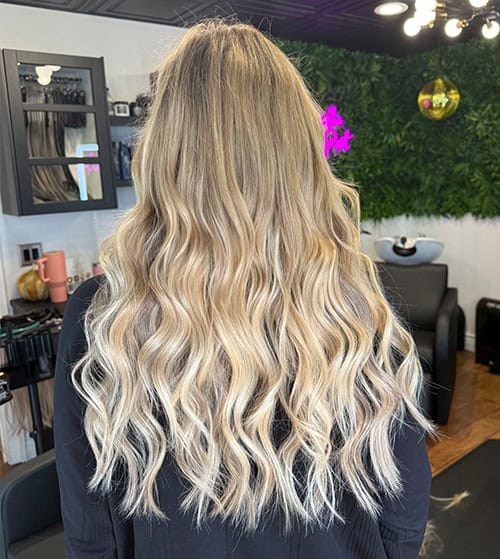 A person with long, wavy blonde hair by Jessie Lu is standing in a salon, facing away from the camera.