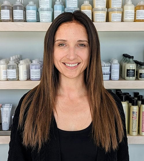 A woman with long straight brown hair stands indoors, smiling at the camera. Shelves with neatly arranged haircare products are visible in the background, highlighting the signature touch of hair by Jessie Lu.