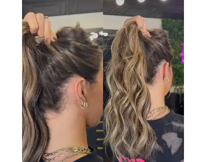 A person with wavy, highlighted hair by Jessie Lu is shown from the back, holding their hair up in a high ponytail with one hand. The person is wearing gold earrings and layered necklaces.