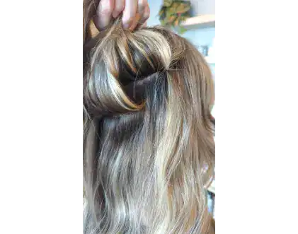 A hand lifts a section of wavy, highlighted blonde hair by Jessie Lu to reveal darker roots underneath.