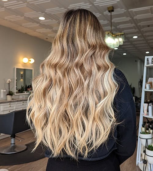A person with long, wavy, blonde balayage hair by Jessie Lu is standing in a modern salon, facing away from the camera.