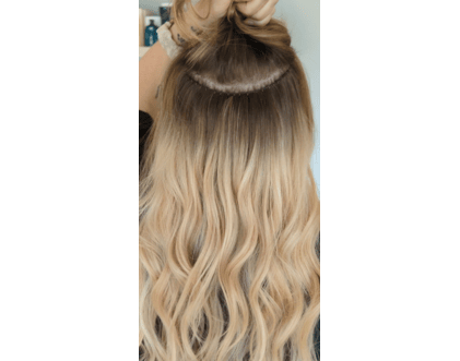 A person holds up a section of long, wavy blonde hair, revealing a row of hair extensions by hair by Jessie Lu attached near the scalp.