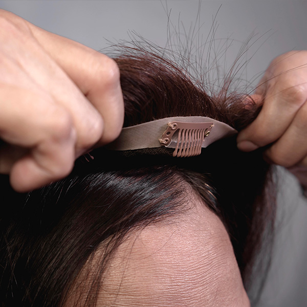 Close-up of hands attaching a hairpiece with a clip to the front hairline on a person's scalp.