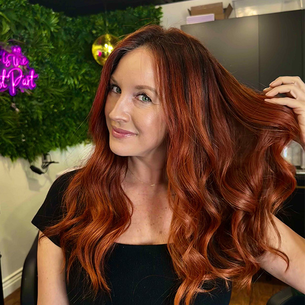 Woman with long, wavy copper-red hair poses indoors, holding a section of her hair. She is wearing a black top, with greenery and salon decor in the background.